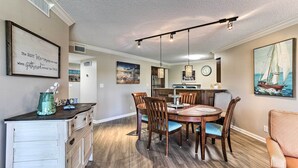 A dining area with a wooden table and chairs, a kitchen in the background, and a painting on the wall.