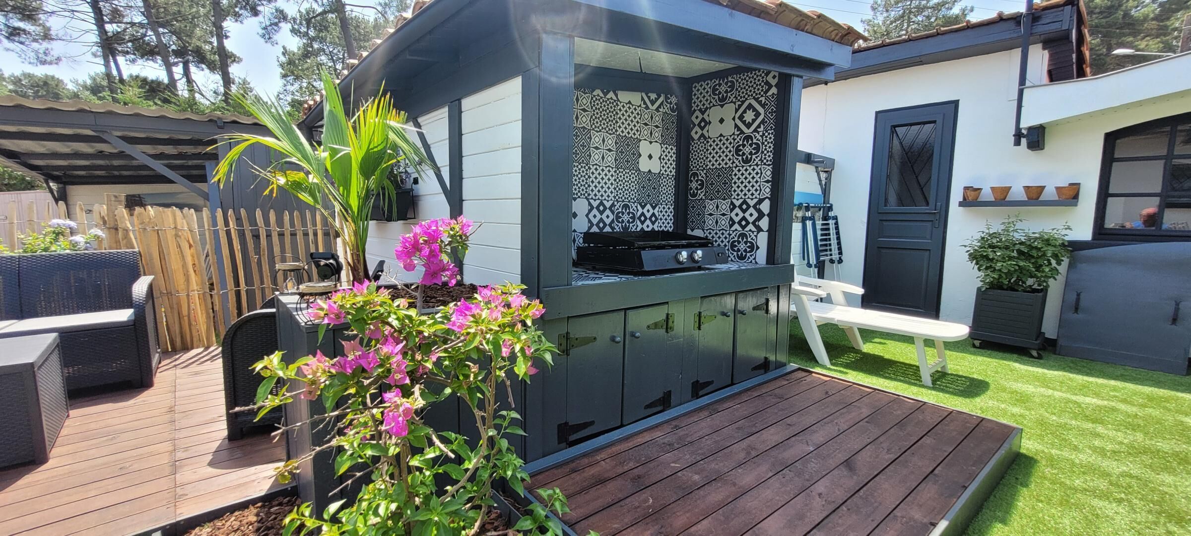 Una cocina exterior moderna con una parrilla empotrada, rodeada por una terraza de madera y una pequeña zona ajardinada con flores rosas.