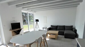 Un salon moderne avec une table à manger blanche, des chaises en bois, un canapé gris et un téléviseur posé sur un meuble en bois.