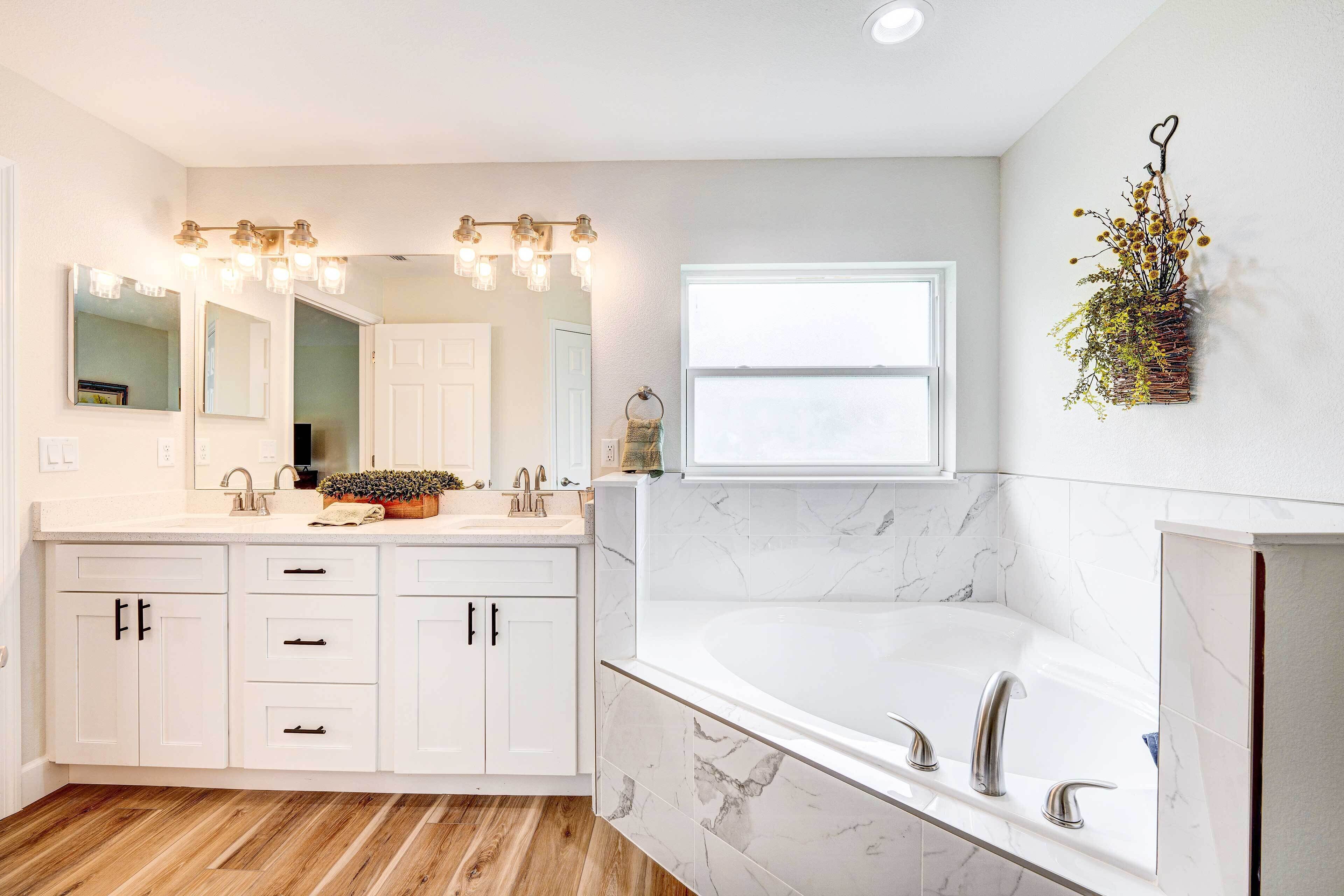 A modern bathroom with a large mirror, a vanity with drawers, a bathtub, and a window.