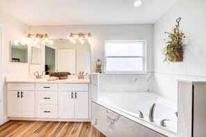 A modern bathroom with a large mirror, a vanity with drawers, a bathtub, and a window.