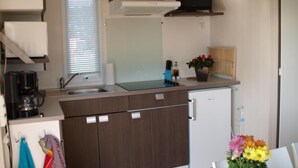 Eine kompakte Küche mit Mikrowelle, Kaffeemaschine und kleinem Kühlschrank. Ein Fenster mit Jalousien, ein Spülbecken und ein kleines Hängeschränkchen mit einem Handtuch.
