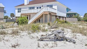 Una casa de dos pisos con balcón, una terraza de madera y una playa arenosa al frente.