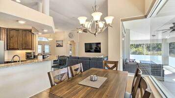 A modern kitchen with wooden cabinets, a dining area with a wooden table and chairs, and a living room with a gray sofa and a TV mounted on the wall.