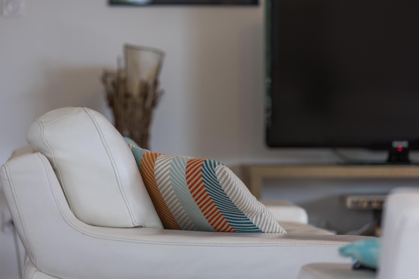 Een witte leren bank met een kussen met een patroon, een flatscreen-televisie en een decoratieve vaas in een woonkamer.