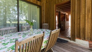 A wooden interior with a dining table set for four, a vase with flowers, and a view of the outdoors through large windows. - Vieux Chêne - Vert - Gîte (VERT)