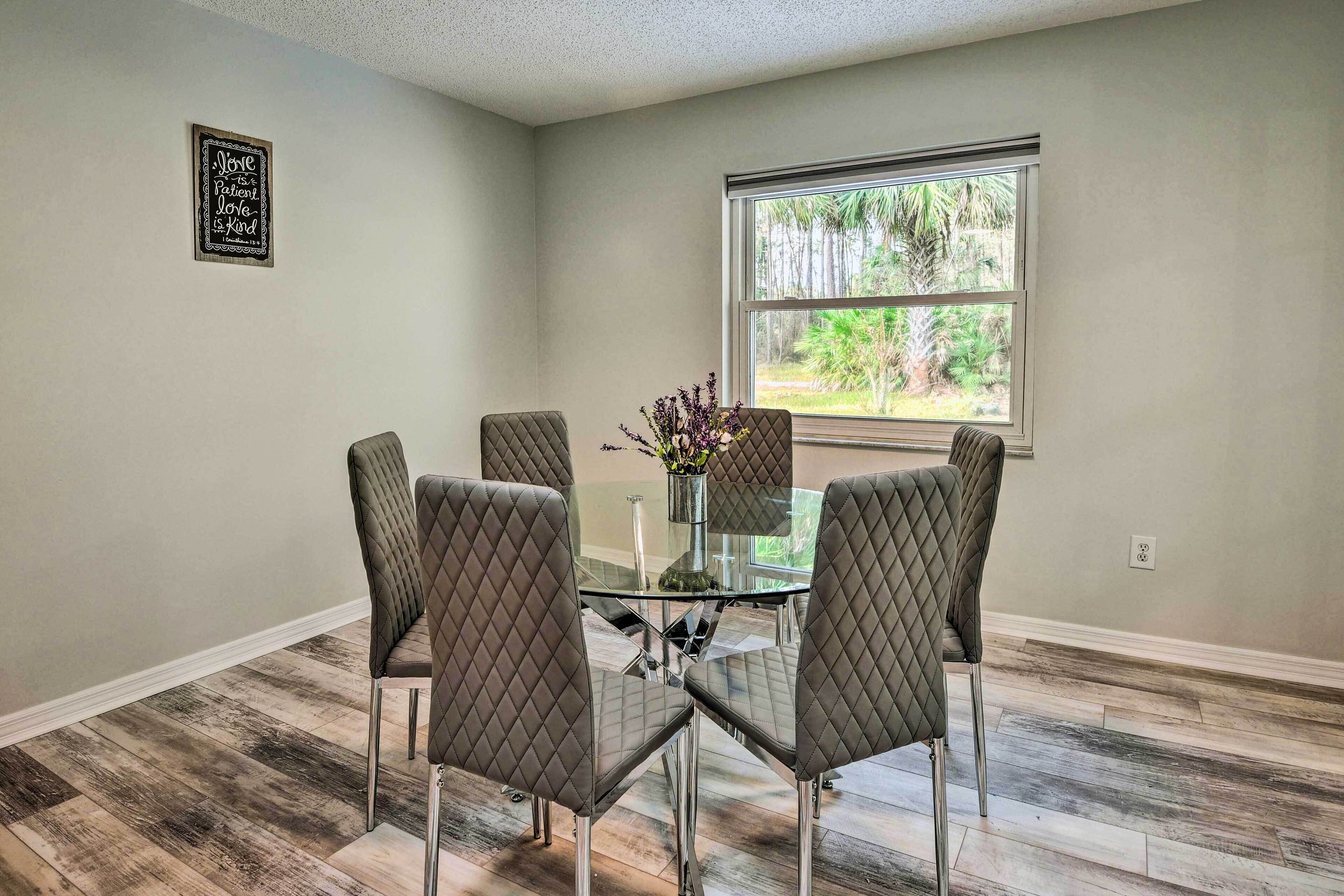 A dining area with a glass table surrounded by four cushioned chairs, a vase with flowers, and a window with a view of trees.