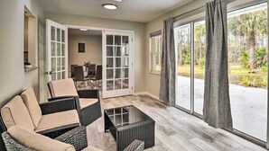Una habitación con mobiliario de mimbre, una mesa de centro de vidrio y vistas a un jardín a través de grandes ventanales.