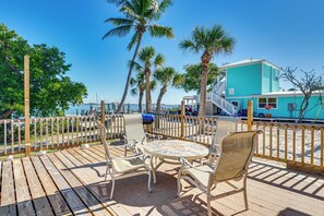 A wooden deck with a round table and chairs, overlooking a beach with palm trees and a turquoise building in the background. - Little Gasparilla Home: Walk to Beaches! (Placida)