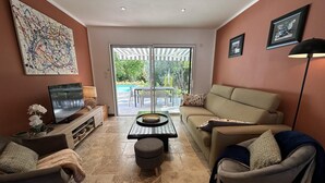 Sala de estar con televisor, sofá, sillones, una mesa de centro y vista a una piscina a través de puertas de vidrio.
