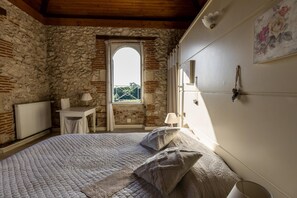 Quarto com parede de pedra, teto de madeira, cama com colcha, uma mesa pequena e janela com vista.