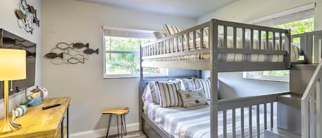 Uma cama beliche com roupa de cama listrada, uma pequena escrivaninha de madeira com abajur e um objeto decorativo na parede.