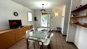Ein Essbereich mit einem Glastisch, weißen Stühlen, einem Holzschrank, einem Fernseher und einer Deckenleuchte.