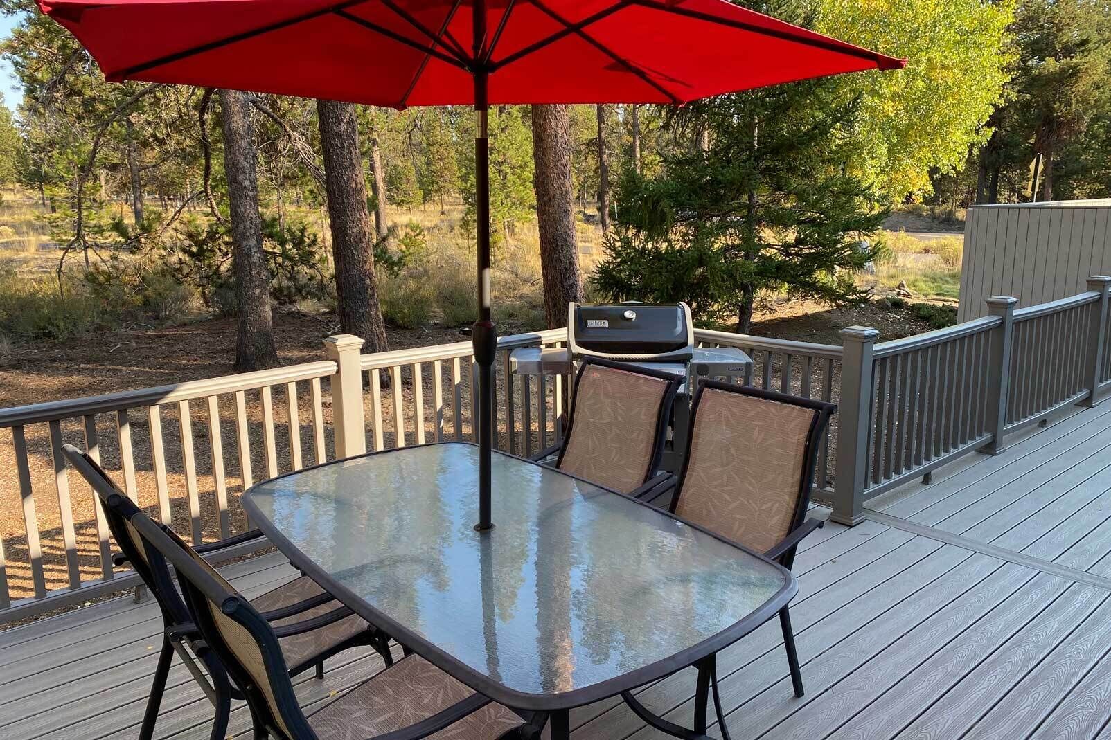A deck with a glass-top table and chairs, a red umbrella, and a grill.
