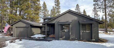 A two-story building with a garage and a covered porch, surrounded by snow and tall trees.