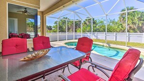 Eine überdachte Terrasse mit einem Glastisch und roten Stühlen, mit Blick auf einen Swimmingpool und üppiges Grün.