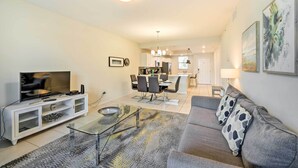 A modern living room with a flat-screen TV, a glass coffee table, and a gray sofa with patterned cushions. A dining area with a white table and chairs is visible in the background. - Resort Getaway w/ Patio: 6 Mi to Disney Parks! (Kissimmee)