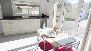Une cuisine lumineuse avec une grande fenêtre, une table à manger avec des coussins rouges et une vue sur un bâtiment jaune à l’extérieur.