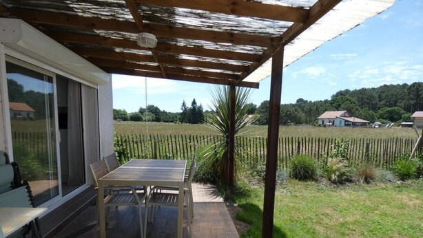 A covered patio with a table and chairs, a sliding glass door, and a view of a vineyard and houses in the background. - Les Palombières - SOUSTONS (SOUSTONS)