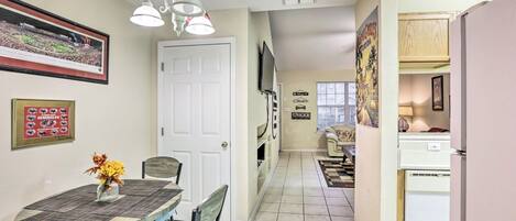 A compact kitchen with a dining area, featuring a white door, a TV mounted on the wall, and a small refrigerator.