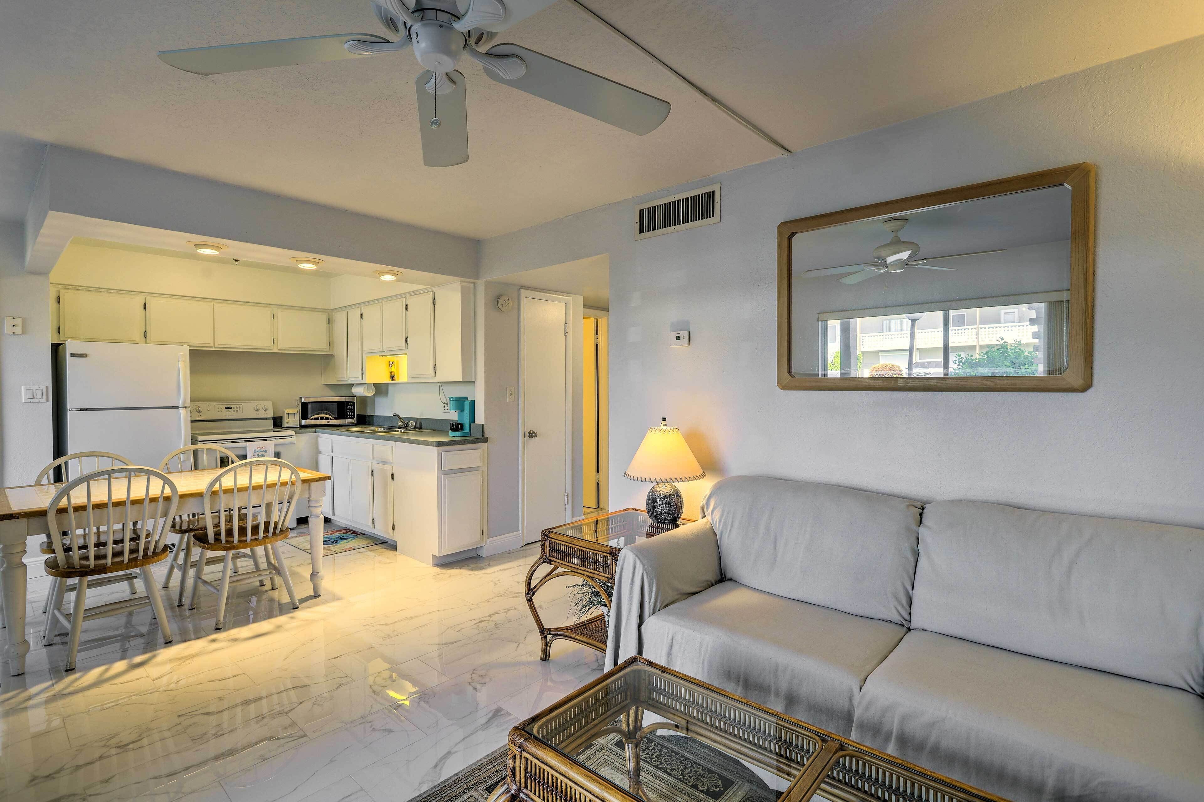 A modern kitchen with white cabinets, a dining area with a table and chairs, a sofa, and a ceiling fan.