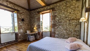 Une chambre avec un mur en pierre, un plancher en bois, un lit recouvert d’une couvre-lit à motifs, un bureau avec une chaise et une fenêtre donnant sur un paysage agréable.