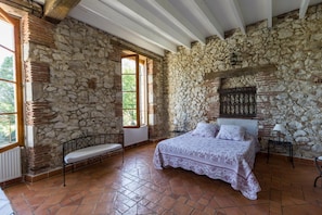 Un dormitorio con paredes de piedra, una cama con colcha estampada, un banco y una ventana.