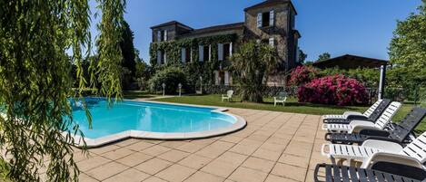 A swimming pool with lounge chairs, a building with a tiled roof, and lush greenery.