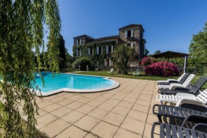 Una piscina con sillas de descanso, un edificio con techo de tejas y exuberante vegetación.