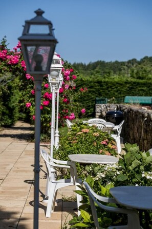 Un patio con sillas y mesas de plástico blanco, un farol blanco y flores rosas al fondo.