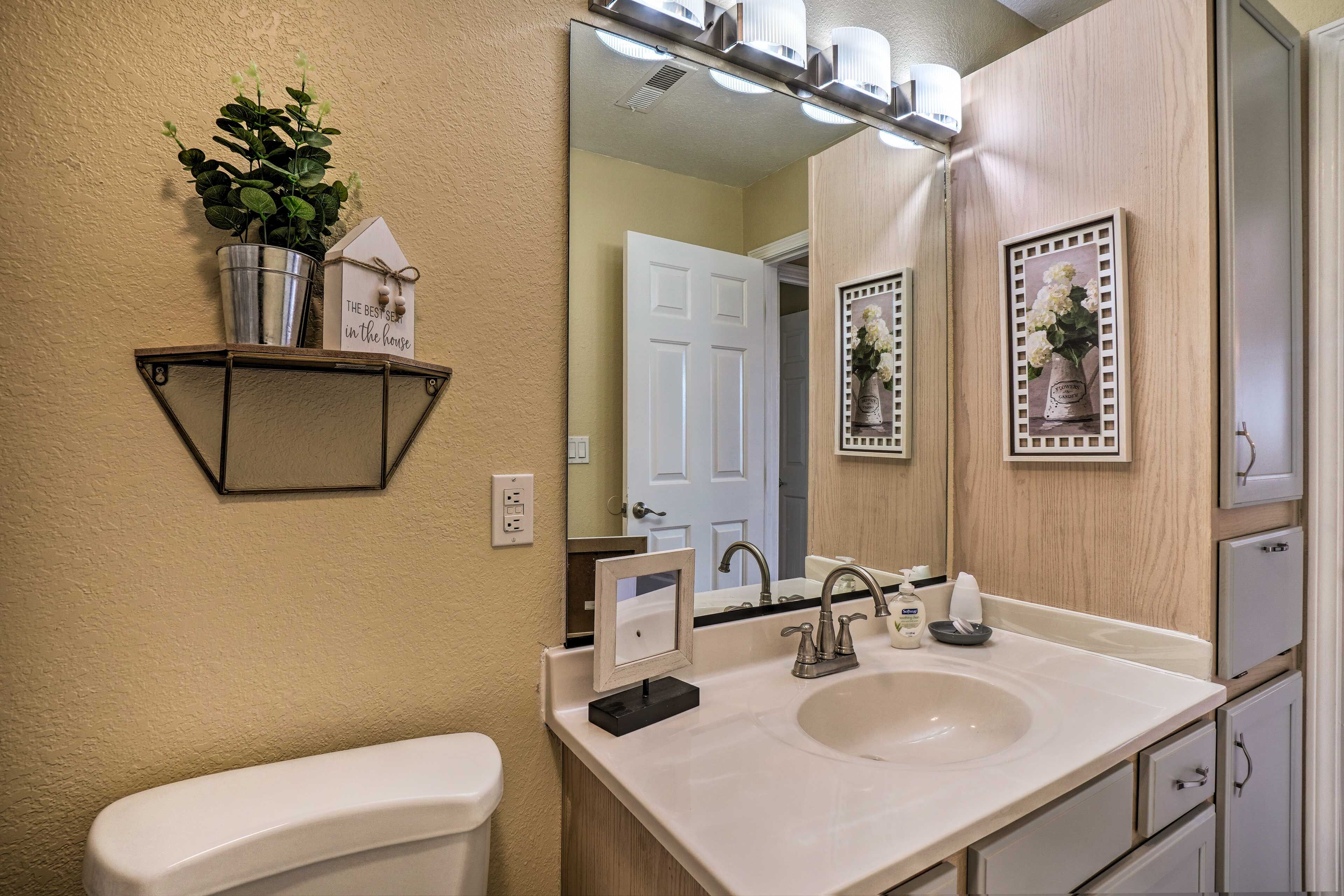 Une salle de bain avec un lavabo blanc, un miroir, des toilettes et une étagère sur laquelle se trouvent une plante en pot et un petit objet décoratif en forme de maison.