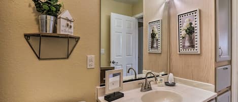 Une salle de bain avec un lavabo blanc, un miroir, des toilettes et une étagère sur laquelle se trouvent une plante en pot et un petit objet décoratif en forme de maison.