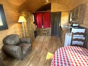 A cozy cabin interior with a wooden floor, a sofa, a table, a lamp, a red curtain, and a kitchenette.