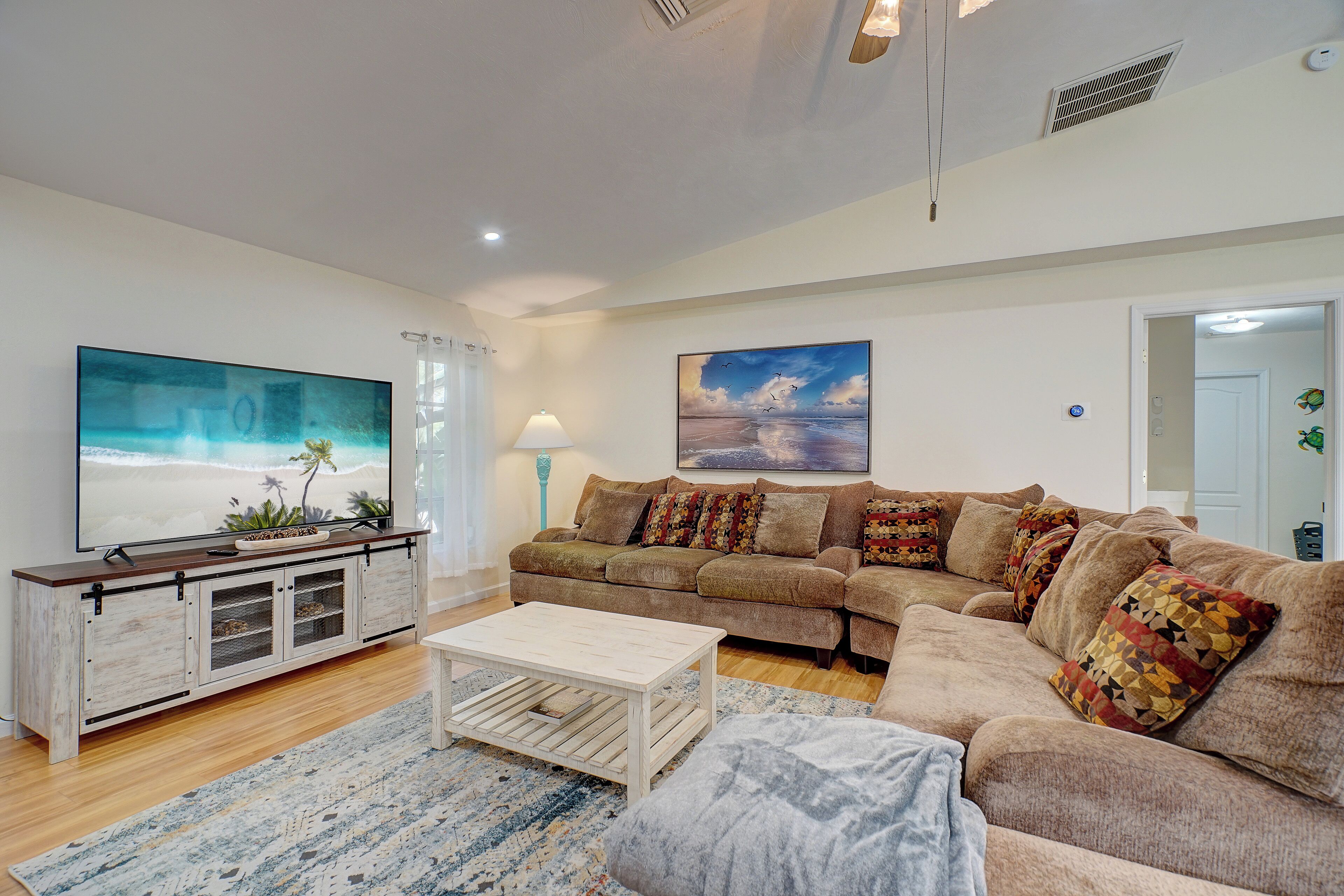 A living room with a large flat-screen TV, a wooden coffee table, a beige sectional sofa with patterned cushions, and a blue patterned rug.