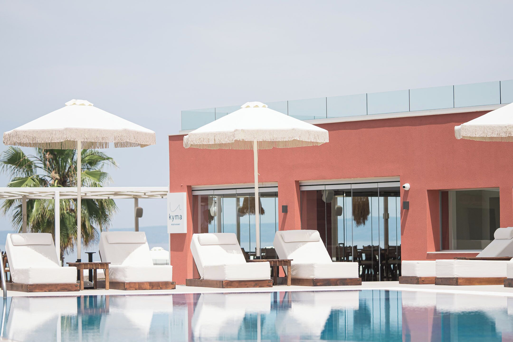 A poolside area with white lounge chairs and umbrellas, a glass balustrade, and a building with large windows.