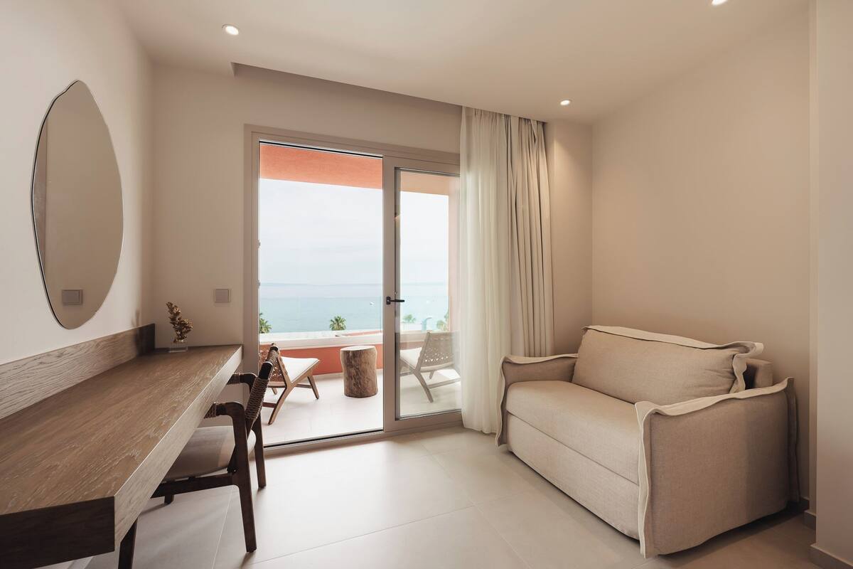 A modern living room with a sofa, a dining table with chairs, a large mirror, and a sliding glass door leading to a balcony with a view of the sea.