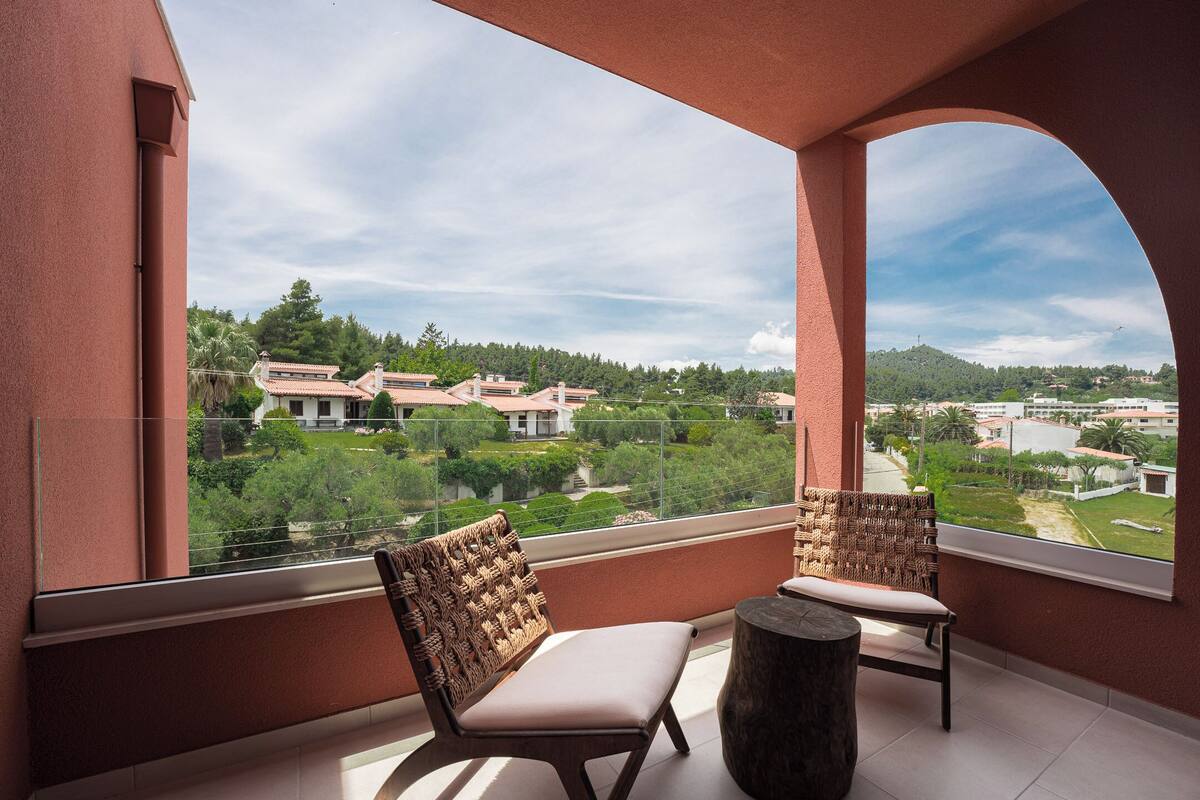 A balcony with two wicker chairs and a wooden table, overlooking a residential area with houses and greenery.