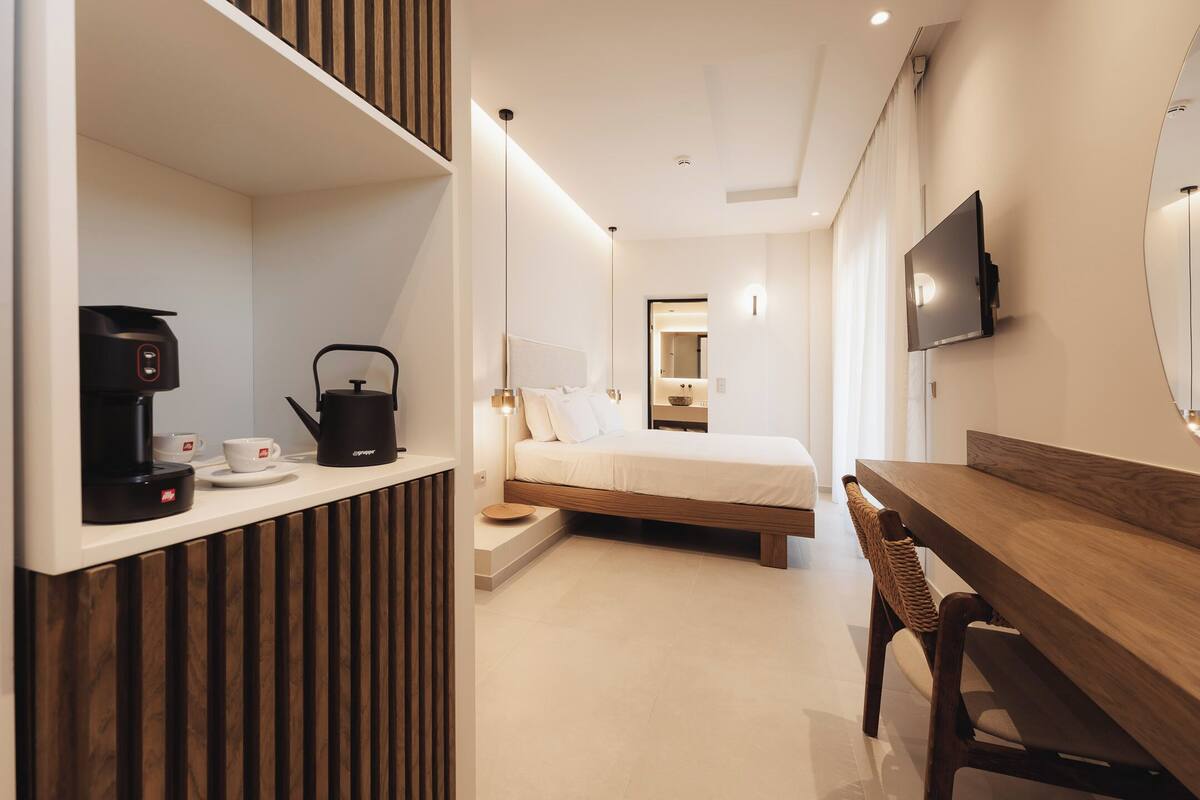 A modern hotel room with a bed, a wooden desk, a coffee machine, and a TV mounted on the wall.