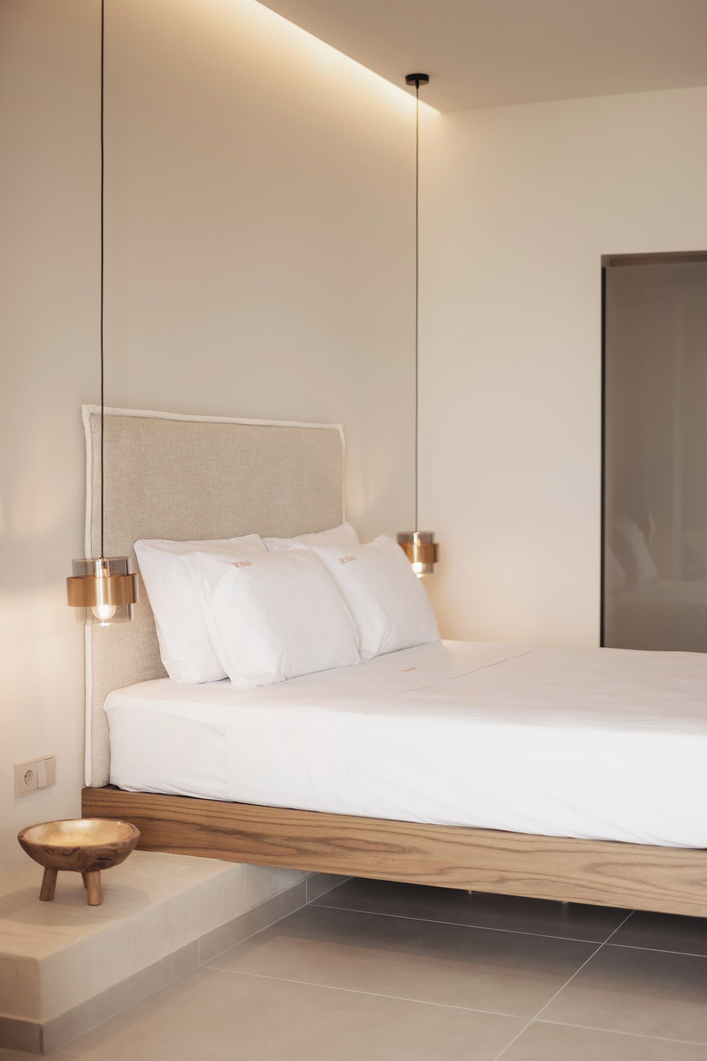 A modern bedroom with a minimalist design, featuring a bed with white bedding, a wooden headboard, and a small wooden stool.