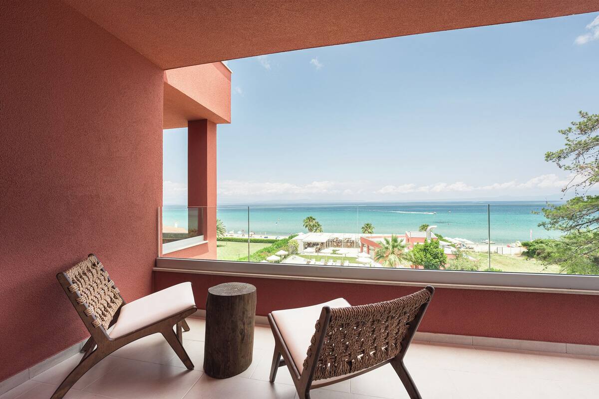A balcony with two wicker chairs, a wooden stool, and a view of the ocean and resort buildings.