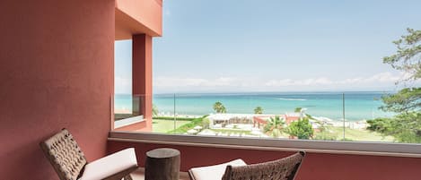 A balcony with two wicker chairs, a wooden stool, and a view of the ocean and resort buildings.