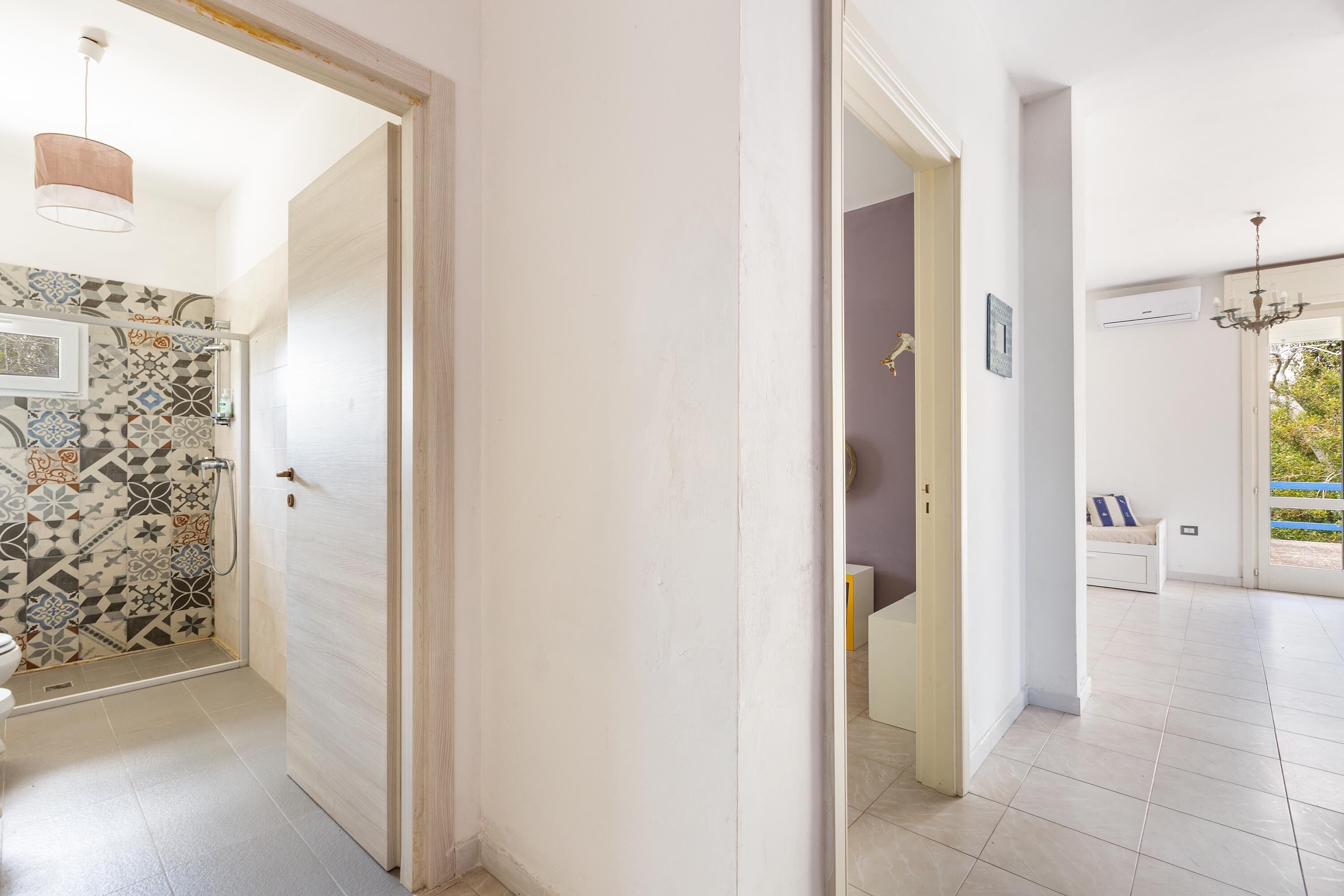 Un pasillo con un papel tapiz estampado, una puerta que da a un baño y vistas a un balcón con barandilla.