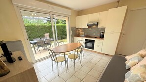 Una cocina moderna con armarios blancos, horno integrado y cafetera. Hay una zona de comedor con mesa y sillas, y un amplio ventanal corredero que da a un patio exterior.