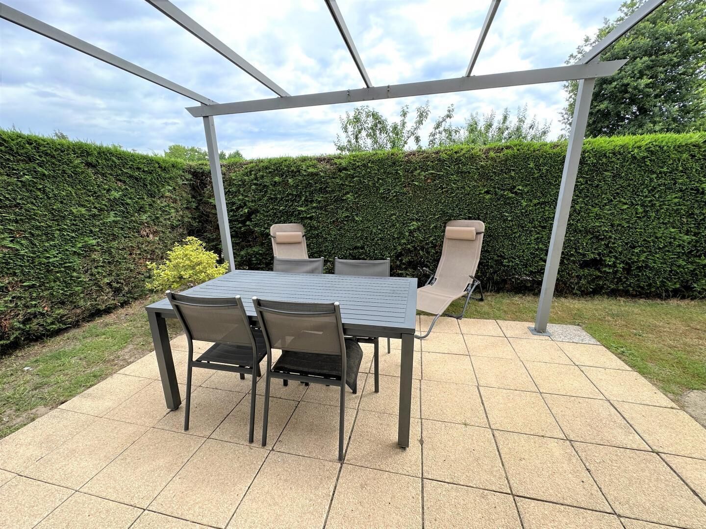 A covered patio with a table and chairs, surrounded by a hedge and a clear sky.
