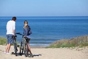 Twee personen staan op een zandstrand met fietsen, en kijken uit over de oceaan.