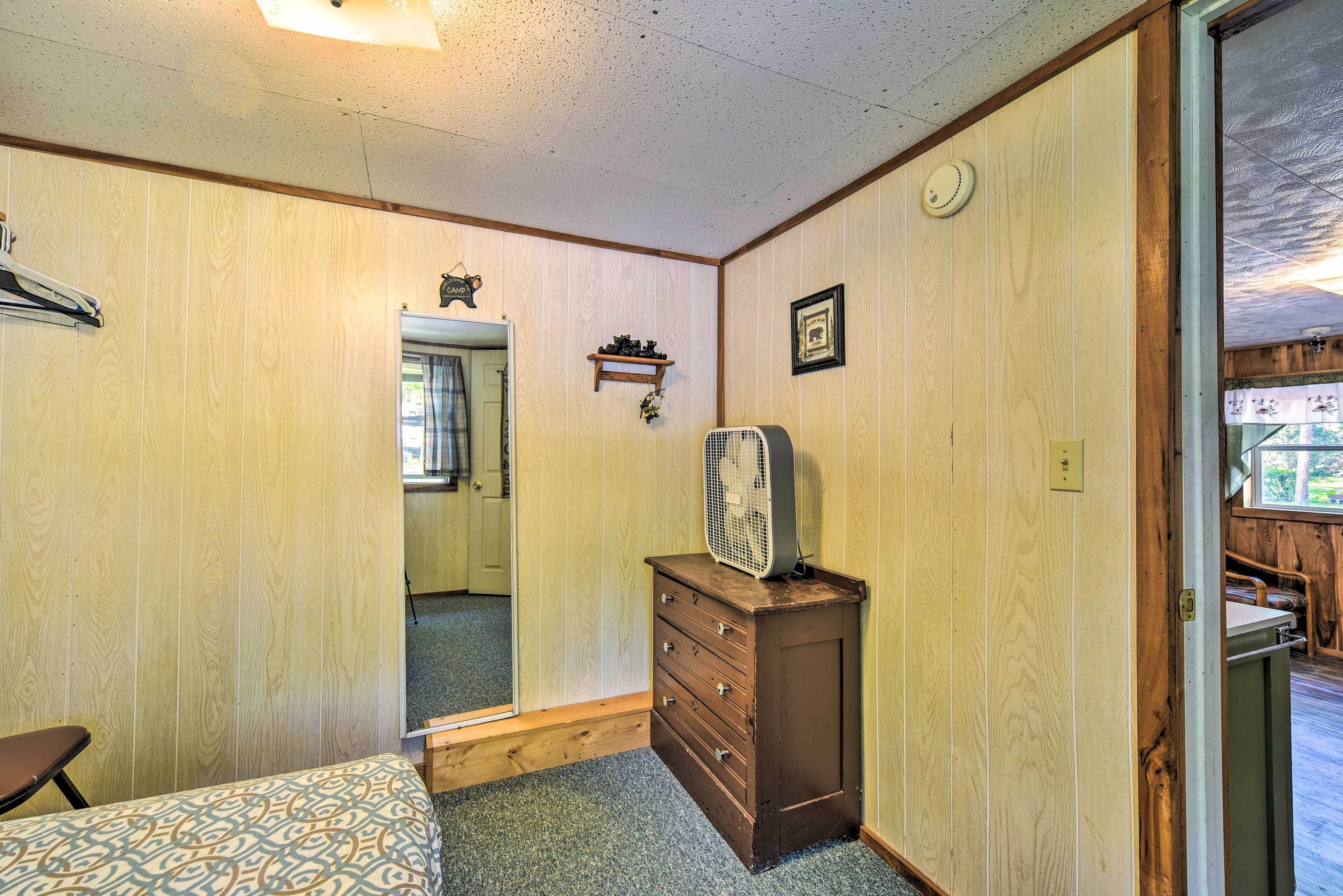 A room with wooden panel walls, a wooden dresser with drawers, a bed with patterned bedding, a fan on the dresser, a mirror, and a door leading to another room.
