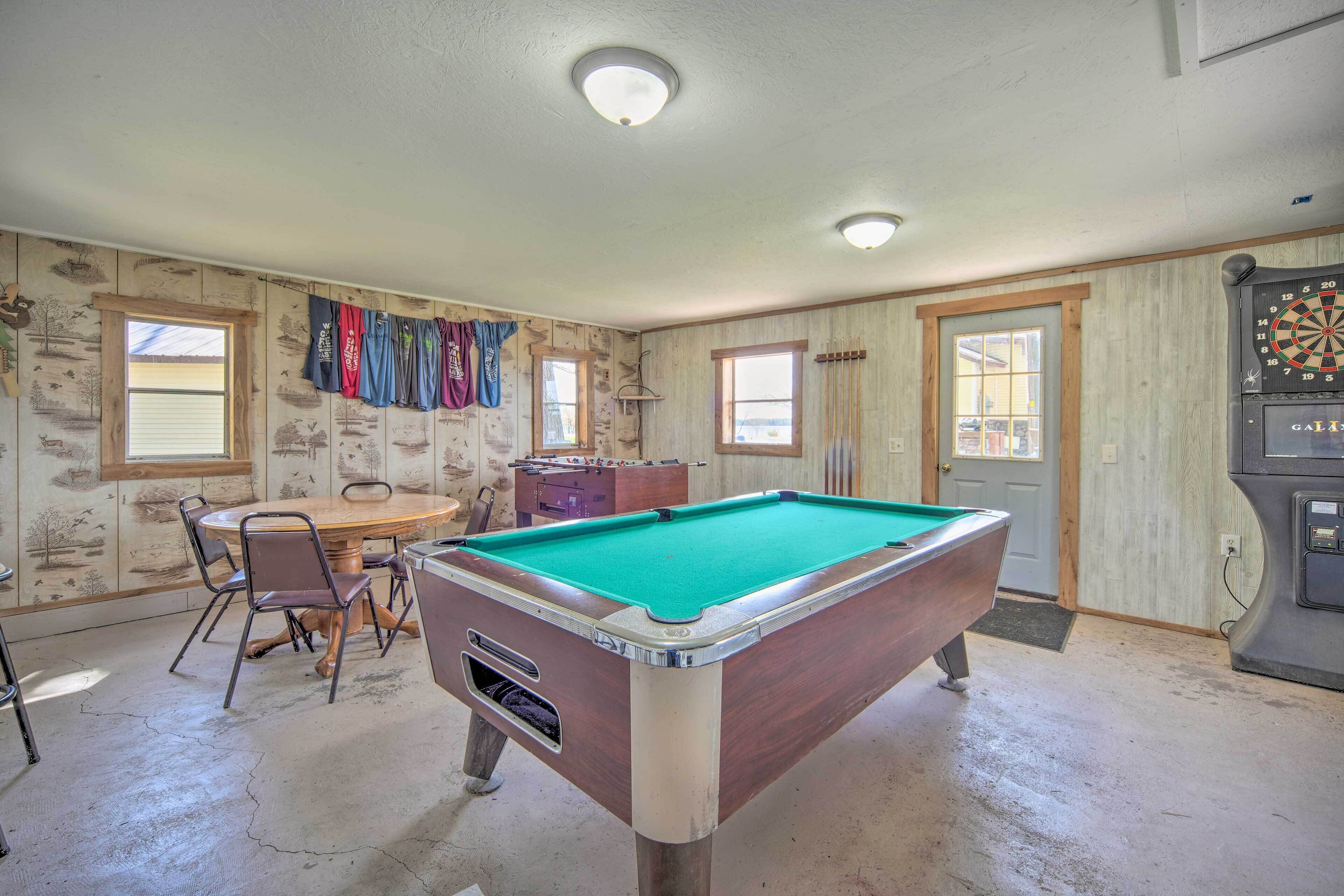A pool table in a room with wooden walls, a dartboard, and a window.