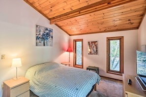 A bedroom with a wooden ceiling, a bed with a patterned bedspread, a nightstand with a lamp, a desk with a computer, and two windows with views of trees.