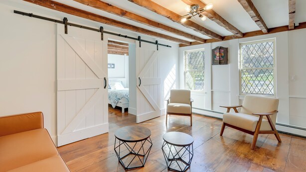 Ein geräumiges Zimmer mit Holzbalken, einer Schiebetür im Bauernhausstil, einem Bett, zwei Stühlen und zwei runden Tischen.