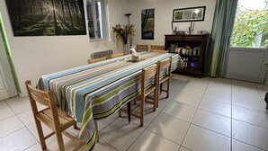 Een eetgedeelte met een geruit tafelkleed, houten stoelen, een radiator, een raam met gordijnen en een boekenkast met diverse spullen.